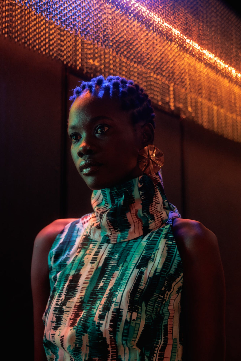 Woman with colorful hair and dress standing in front of a decorative wall at night.