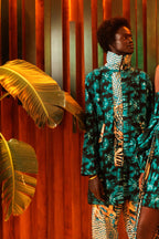 Two women in vibrant, patterned outfits standing against a warm-toned wall with plants.