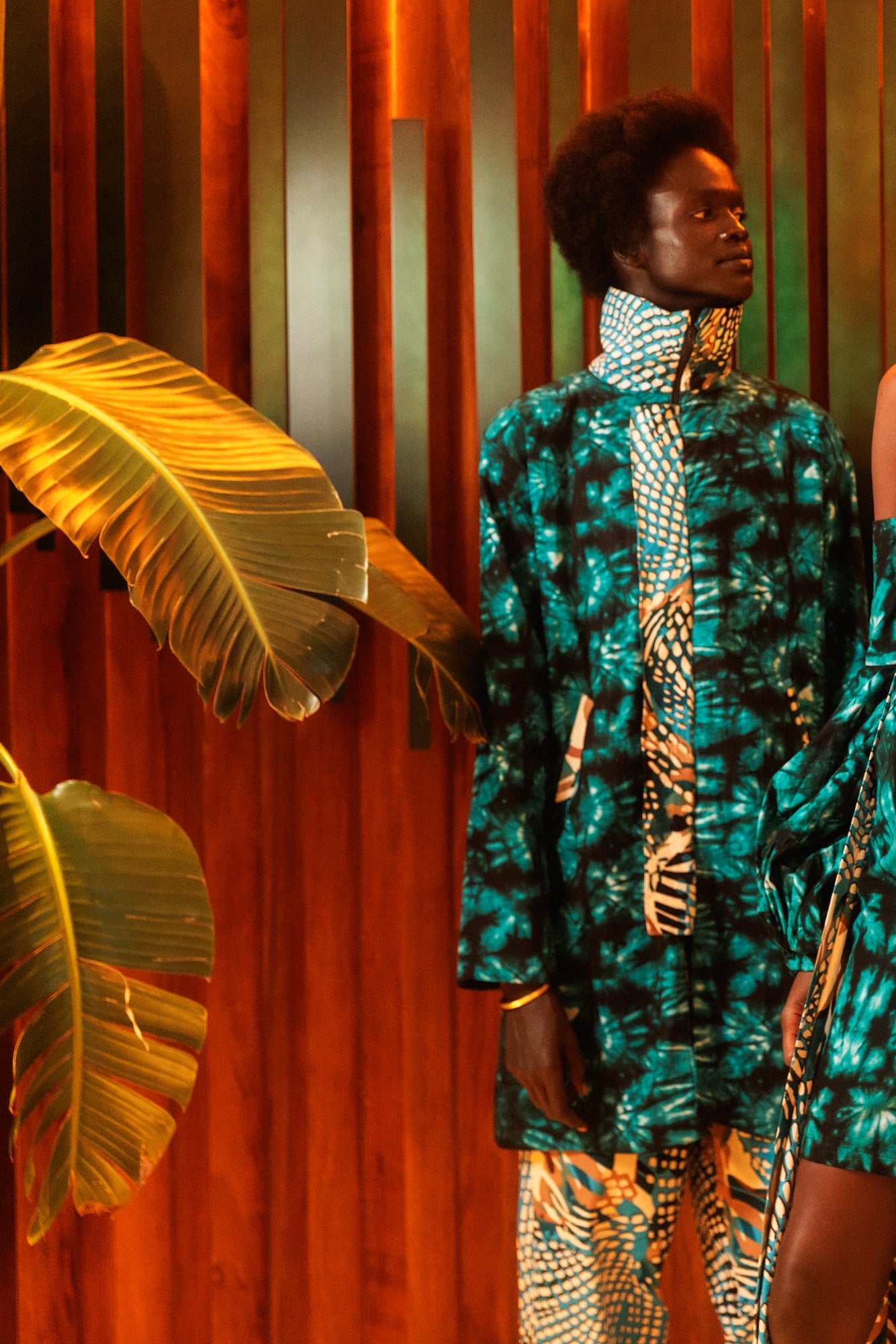 Two women in vibrant, patterned outfits standing against a warm-toned wall with plants.