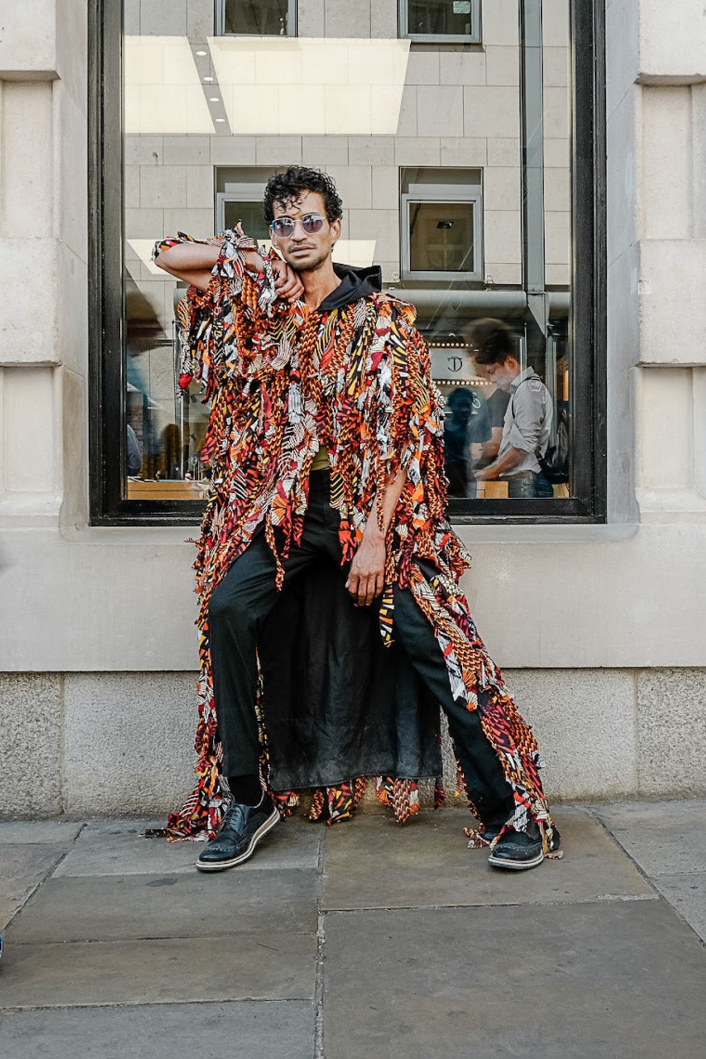 Man wearing a patterned shirt and sunglasses standing in front of a building with a window display.